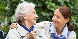 A caregiver and an elderly woman are enjoying a moment together outdoors, with the woman holding a cup of tea.