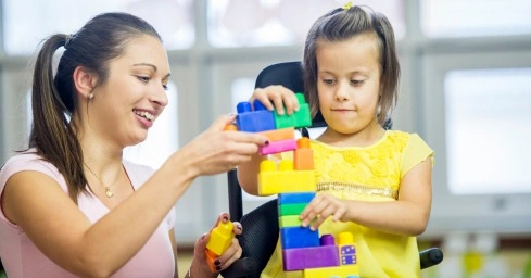 A caregiver is helping a young child in a wheelchair build a structure with colorful blocks, creating an engaging learning activity.