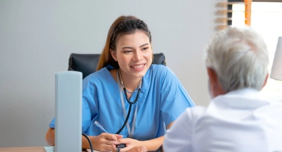 A healthcare professional in blue scrubs smiling and engaging in a conversation with an elderly male patient, as they discuss medical matters in a clinic setting.