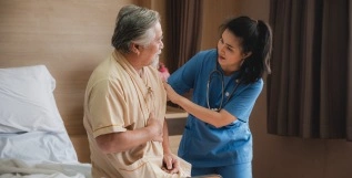 A healthcare worker is assisting an elderly man in a hospital bed, offering support and care with a compassionate expression.