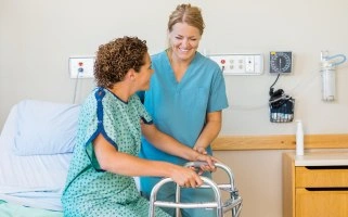  This image shows a nurse, dressed in green scrubs, assisting a patient who is using a walker to stand or move.