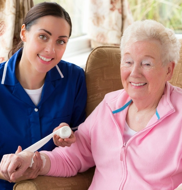 A caregiver in a blue uniform smiling and assisting an elderly woman with bandaging her wrist