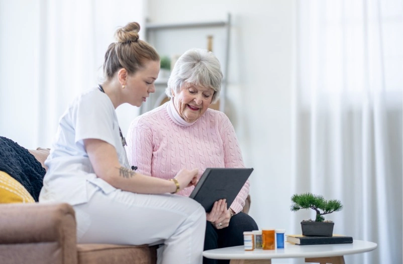 A caregiver in blue scrubs pushing a wheelchair with an elderly man sitting in it, as they engage in a conversation.