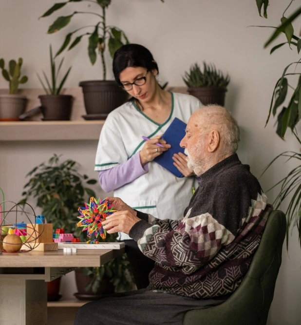 A caregiver in a white uniform, holding a notebook and standing behind an elderly man engaged in an activity with colorful toys at a home setting.