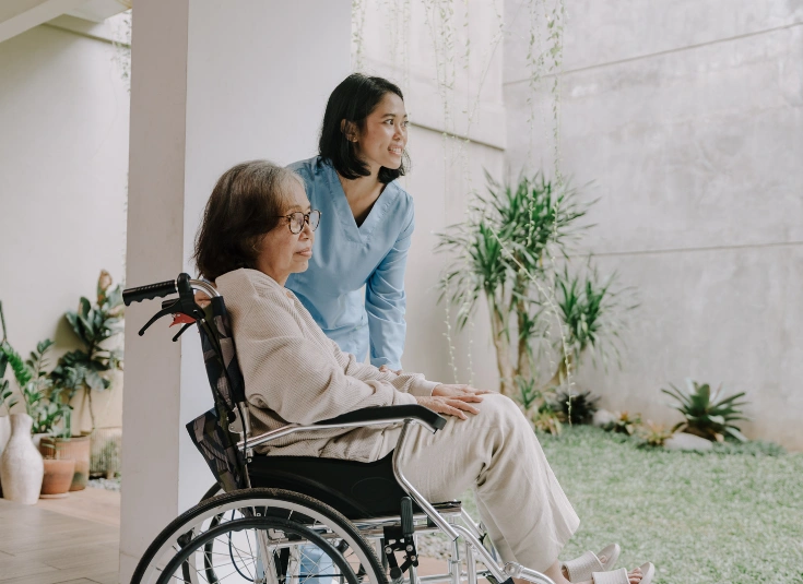 A caregiver in a light blue medical uniform pushing an elderly woman in a wheelchair outside, with plants and greenery in the background.