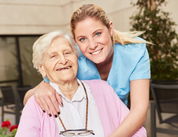 A happy elderly woman sitting in a chair with her caregiver, a young woman wearing a blue medical uniform, who is smiling and hugging her from behind.