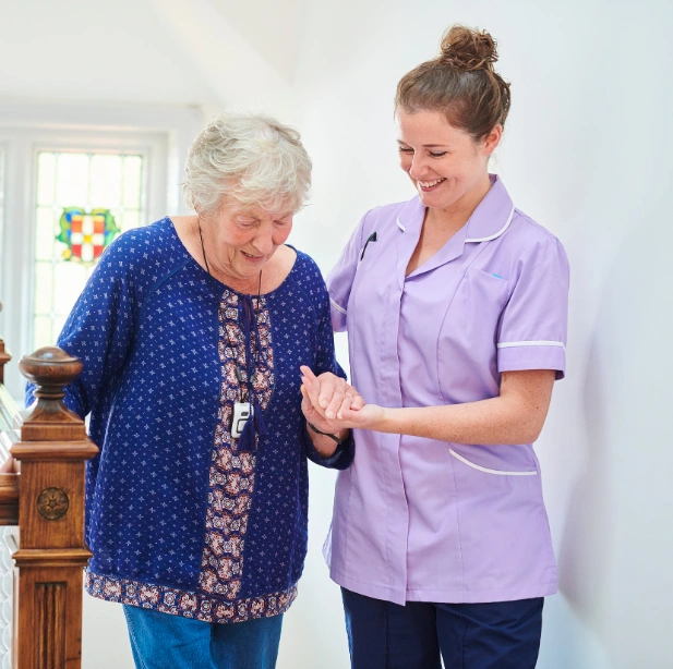 A caregiver in purple scrubs assisting an elderly woman as they walk together in a hallway, with a warm, supportive interaction.
