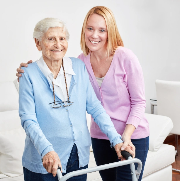 A caregiver with red hair in a pink cardigan stands next to an elderly woman using a walker, both smiling at the camera in a home setting
