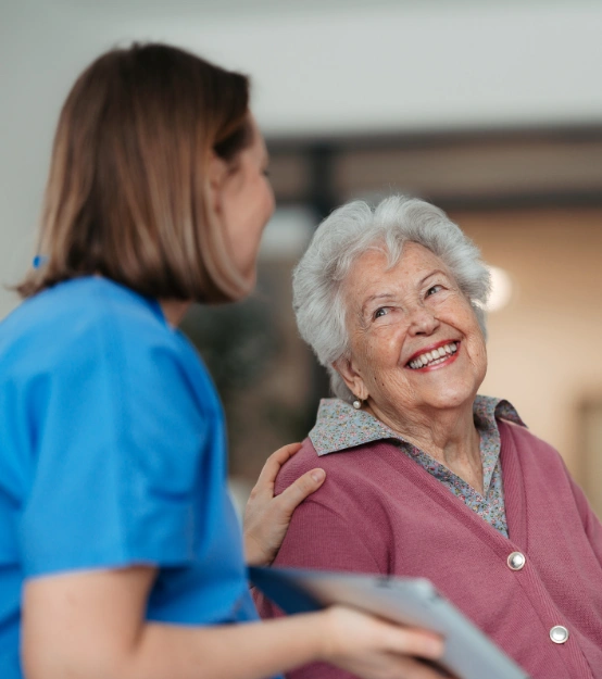 A caregiver in blue scrubs smiling and engaging with an elderly woman in a pink sweater, showing a positive interaction in a healthcare setting.