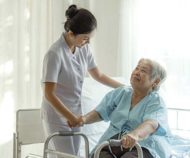 A caregiver in a light gray uniform assisting an elderly woman in a hospital gown to stand up using a walker.