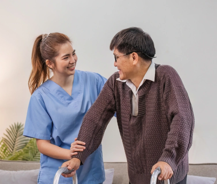 A caregiver in a blue medical uniform helping an elderly man with a walker while both are smiling at each other in a living room setting.