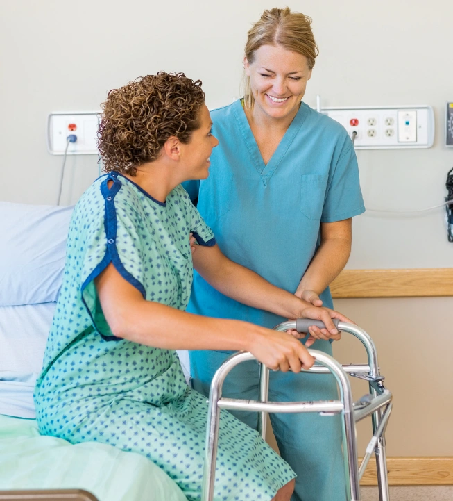 A caregiver in green scrubs assisting a woman in a hospital gown with a walker, both smiling at each other in a hospital room.